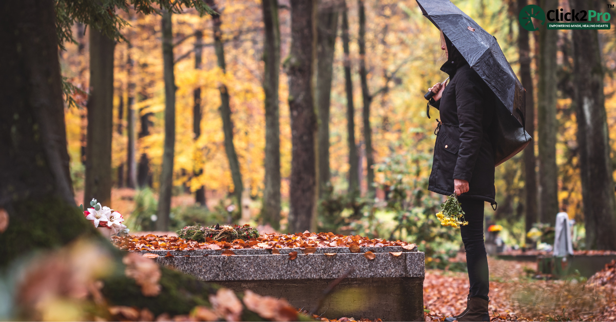 Person grieving at a gravesite during autumn, holding flowers and umbrella, reflecting on loss.